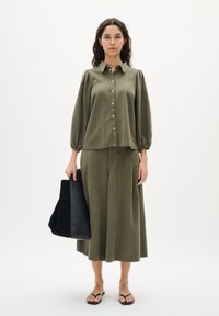 Olive roheline, laia lõikega särk nööpidega ja põlvini puhviku varrukad, kombineeritud vastava laiade seadmete pükstega. Must käekott, lamedad sandaalid.