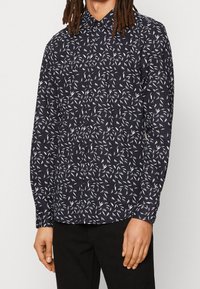 Laivaston napillinen paita, jossa on valkoisia lehtikuviota, pitkät hihat ja kaulus. Valmistettu kevyestä kankaasta, jossa on normaali istuvuus ja hihansuut.