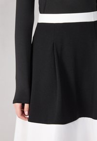 Robe noire et blanche avec un corsage noir ajusté et une jupe blanche évasée, accentuée par une large ceinture blanche. Tissu lisse et texturé.