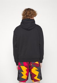 Persona con capelli ricci che indossa una felpa con cappuccio nera e pantaloncini colorati con motivo a zigzag, vista di spalle su uno sfondo bianco uniforme.