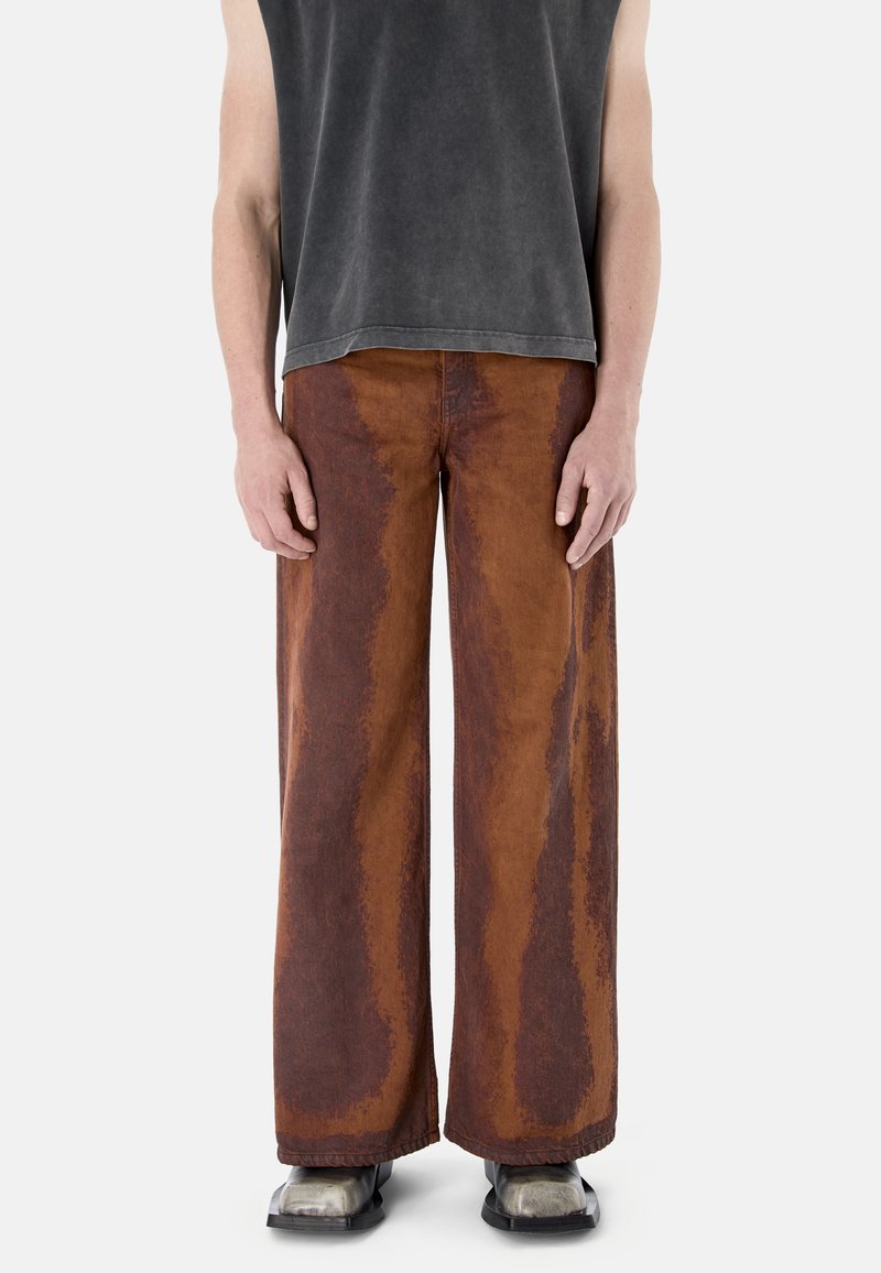 Person trägt weit geschnittene braune und rostfarbene Hosen, ein ärmelloses dunkelgraues Oberteil und silberne Schuhe mit quadratischer Spitze, steht vor weißem Hintergrund.