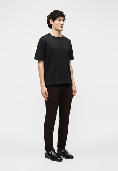 Νεαρός άνδρας όρθιος, φορά απλό μαύρο κοντομάνικο μπλουζάκι, σκούρο στενό παντελόνι και μαύρα δερμάτινα μοκασίνια, με φόντο ανοιχτό γκρι.