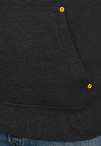 Haut à capuche noir avec une poche avant, comportant un bouton d'accent jaune et une texture côtelée au niveau de l'ourlet. Le matériau semble doux et extensible.