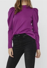 Figurbetontes, langärmliges Oberteil in Lila aus strukturiertem Stoff mit gepufften Schultern, kombiniert mit hochgeschnittenen schwarzen Jeans.