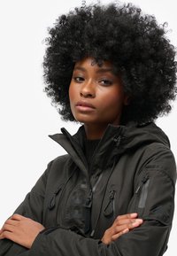 Schwarze isolierte Jacke mit hohem Kragen, mehreren Reißverschlusstaschen und Schnürdetails. Glatte Stoffbeschaffenheit und eine moderne Passform.