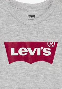 Pilkas megztinis su apykakle, kuriame yra didelis raudonas Levi's logotipas su baltu tekstu, esantis krūtinės centre, ir mažesnis etiketas apykaklės viduje.
