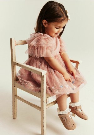 Jeune fille en robe rose à volants et chaussettes en dentelle, assise sur une chaise en bois rustique, regardant vers le bas et souriant doucement.