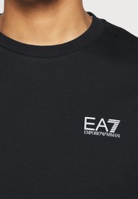 T-shirt nera in cotone con scollo rotondo. Logo bianco ricamato sul petto a sinistra. Tessuto morbido, vestibilità classica, design minimalista.