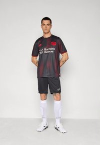 Črno-rdeč nogometni dres iz mrežaste tkanine z ustvarjalno prelivno zasnovo, na katerem so beli logotipi, kombiniran s črnimi kratkimi hlačami, belimi nogavicami in kopačkami.