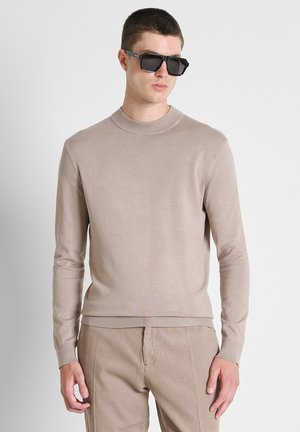 Junger Mann mit dunkler Sonnenbrille, beigem langärmligem Pullover und beigen gerippten Hosen, stehend vor einem schlichten hellen Hintergrund.