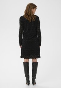 Rochie din dantelă neagră cu mâneci lungi și tiv ondulat. Modelul poartă cizme negre până la genunchi. Materialul neted și texturat îmbunătățește designul.