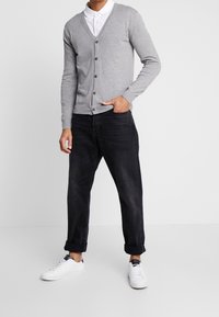 Cardigan grigio lavorato a maglia con scollo a V, chiusura con bottoni, abbinato a jeans scuri e sneakers bianche, con maniche arrotolate e accenti a contrasto.