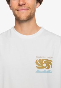 White cotton t-shirt featuring a gold swirl graphic and light blue wave design, branded with "Quiksilver" in a bold font on the chest.