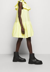 Personne portant une robe jaune ample sans manches et des bottines noires épaisses, debout devant un fond clair uni.