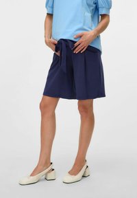 Jūras spēku šorti ar aukliņu vidukļa detaļu, saskaņoti ar gaiši zilu topu. Modelis valkā baltus slingback apavus ar blokveida papēdi.