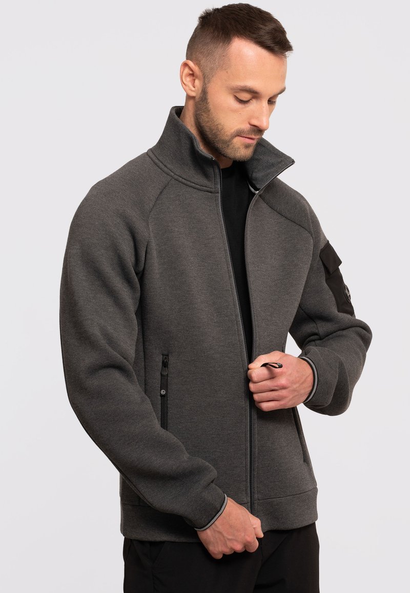 Szara kurtka zapinana na zamek z wysokim kołnierzem, czarnymi akcentami, przednimi kieszeniami zapinanymi na zamek oraz ściągaczami na rękawach. Wykonana z miękkiego, teksturowanego materiału.