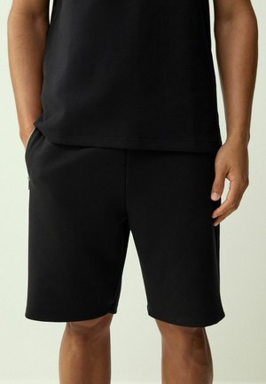 Férfi fekete hétköznapi rövidnadrágban és fekete pólóban, egyik kezét a zsebében tartva, a másik kezét lazán oldalt lógatva áll.