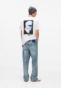 Balta kokvilnas t-krekls ar krāsainu sejas grafiku uz muguras, apvienots ar gaiši ziliem brīvi pieguļošiem džinsiem un pelēku beanie cepuri.