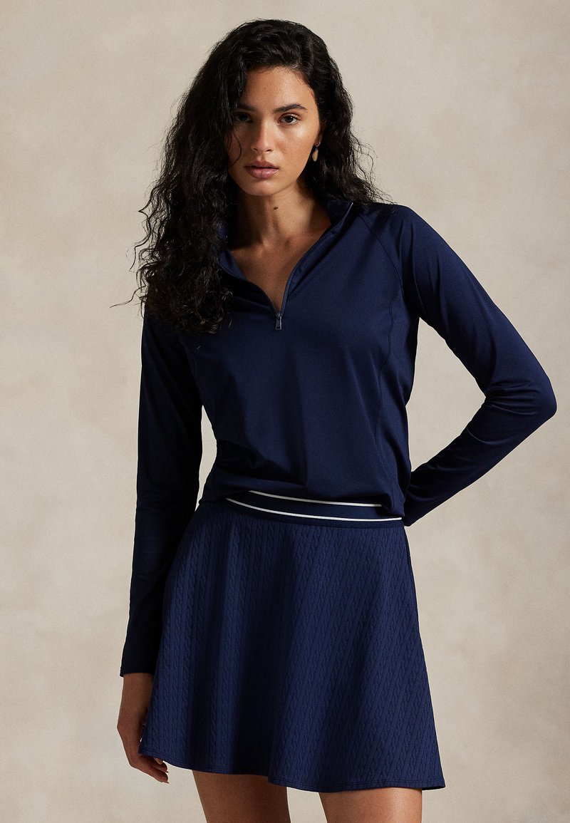 Kvinde med krøllet mørkt hår iført en marineblå langærmet zip-up top og tekstureret navy nederdel med stribet taljebånd står mod en ensfarvet baggrund.