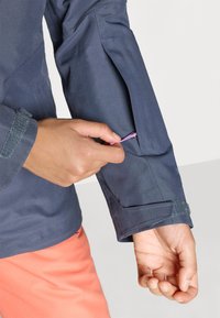 Ναυτική ζακέτα με υφή, που παρουσιάζει μια ασφαλή τσέπη με φερμουάρ στον βραχίονα και λεπτομέρεια με βέλκρο στον καρπό. Ροζ ύφασμα ορατό από κάτω.