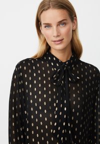 Schwarze durchsichtige Bluse mit goldenen Punktmuster, verfügt über ein Halsband-Design und eine Knopfleiste, die dem Kleidungsstück Textur und Detail verleihen.