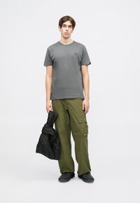 T-shirt cinza de mangas curtas, calças cargo verdes-oliva com bolsos, e uma jaqueta acolchoada preta segurada à mão, acompanhada de sapatos pretos.