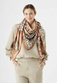 Grand écharpe avec une base beige ornée de motifs de léopard et de feuilles marron, bordée d'une couture orange vif, drapée autour des épaules.