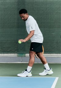 Tennis Spieler trägt ein weißes kurzärmliges Shirt und schwarze Shorts, hält einen grünen Ball. Weiße und hellgrüne Sportschuhe und weiße Socken.