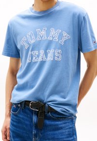 Camiseta de algodón azul claro con el texto "TOMMY JEANS" en blanco, con una tipografía redonda. Combinada con jeans azul oscuro y un cinturón negro trenzado.