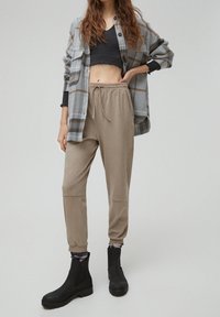 En kvinna står i en grårutigt skjorta, svart crop top, beige joggingbyxor och svarta ankelskor, vilket ger ett exempel på en avslappnad och trendig outfit.