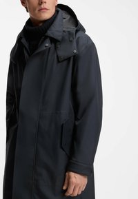 Cappotto blu scuro resistente all'acqua con cappuccio, dotato di bottoni a scatto, tasche laterali e una texture liscia. Adatto per il clima più fresco.