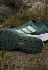 Scarpe da atletica verdi con strisce bianche; suola in gomma testurizzata con un motivo a zigzag; presenta dettagli in blu e arancione.