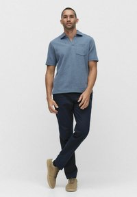 Blå kortärmad polo med bröstficka, matchat med marinblå byxor och ljusbruna sneakers, visad mot en neutral bakgrund.