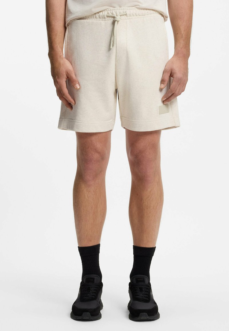 Homme portant un short beige clair avec cordon, des chaussettes noires et des baskets noires, debout devant un fond uni.