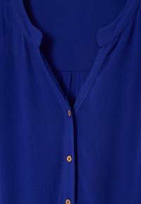 Blusa blu royal con scollatura a V, tessuto textured e bottoni in legno frontali, con pieghe sotto la linea dei bottoni.