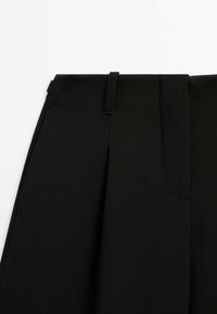 Pantalon noir cintré avec passants de ceinture et pli devant sur un fond clair uni.