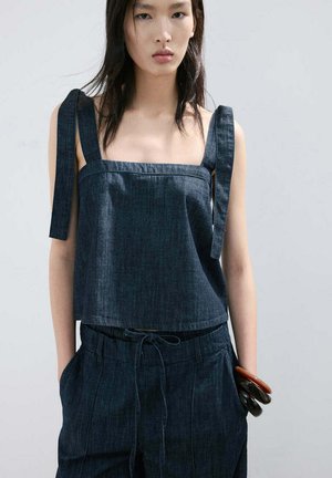 Młoda kobieta ubrana w ciemny dżinsowy top bez rękawów z szerokimi ramiączkami oraz pasujące spodnie, ręce w kieszeniach, na jednolitym jasnym tle.