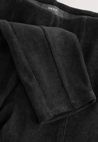 Tissu en maille côtelée noire avec couture visible et détail du poignet, montrant la zone du col avec une étiquette de marque "NEXT".