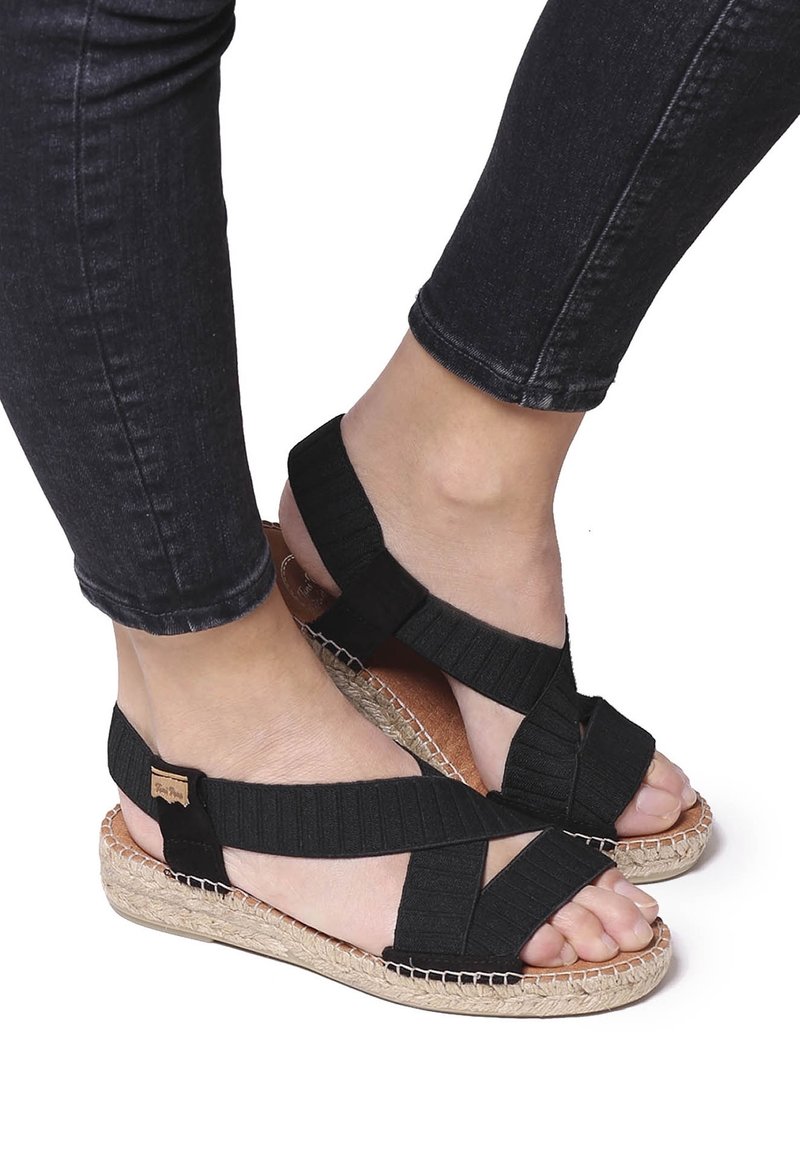 Pieds portant des sandales noires à brides élastiques avec des semelles tissées, associées à un jean noir ajusté.