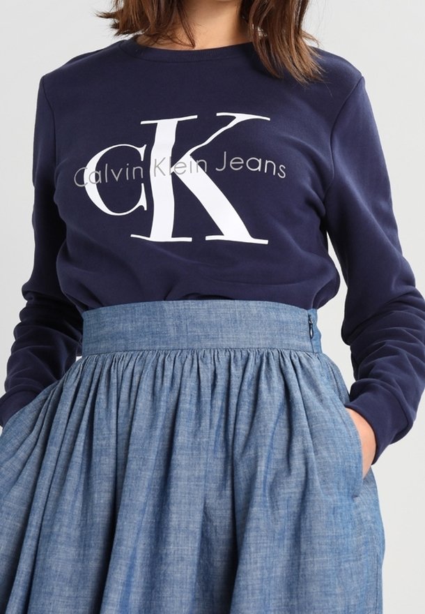 Marinblå sweatshirts med lång ärm med vit "CK"-logotyp och texten "Calvin Klein Jeans"; kombinerad med en ljusblå, högmidjad, plisserad jeanskjol.