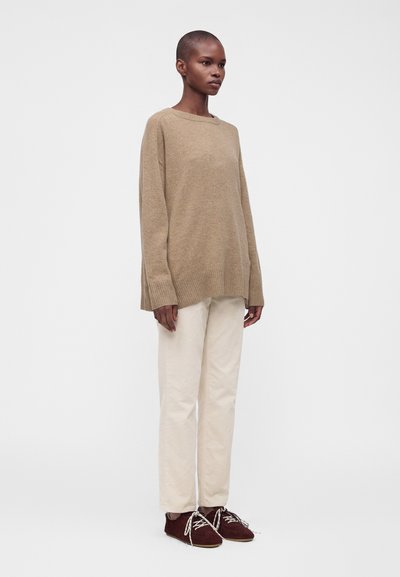 Personne grande aux cheveux courts portant un pull beige ample, un pantalon crème et des chaussures à lacets marron foncé, debout devant un fond clair uni.