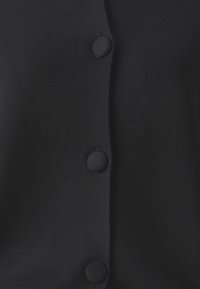 Musta neulottu neuletakki, jossa V-pääntie, kuusi nappikiinnitystä ja sileä, pehmeä rakenne. Kangas vaikuttaa kevyeltä ja hengittävältä.