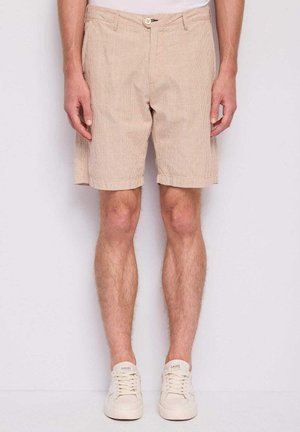 Uomo con pantaloncini in ciniglia beige e scarpe da ginnastica bianche, in piedi contro uno sfondo chiaro e semplice.