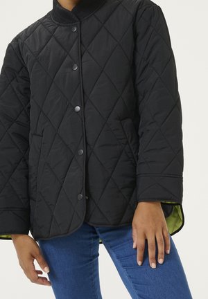 Henkilö, joka pukeutunut mustaan tikattuun takkiin, jossa nepparipainikkeet, näkyvissä hihat ja siniset farkut, kädet rentona sivuilla.