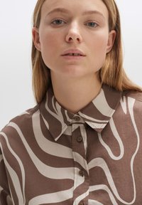 Brown button-up shirt with a contrasting cream wavy pattern, featuring a pointed collar and textured fabric. The buttons are brown.