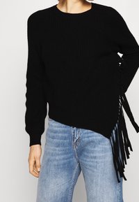 Sort strikket sweater med ribbet tekstur, med asymmetrisk kant og frynse detaljer på den ene side. Kombineret med lyseblå jeans.