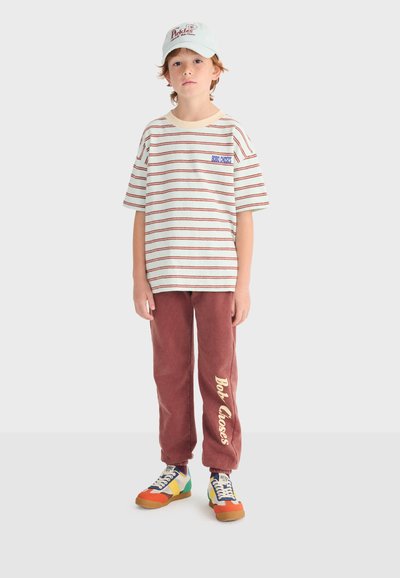 Niño con camiseta de rayas blancas y rojas, pantalones deportivos granate con texto "Bob Closes", zapatillas multicolor y una gorra blanca con texto rojo.