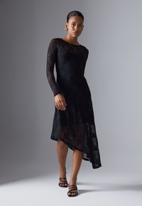 Robe en dentelle noire à manches longues, ourlet asymétrique, couche inférieure transparente et motif floral. Portée avec des sandales noires à lanières.
