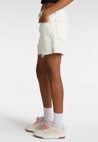 Shorts en denim blanc avec un ourlet effiloché, des poches avant et arrière, et une couture contrastante ; portés avec des chaussures claires et des chaussettes blanches.