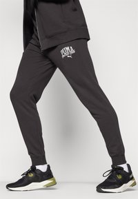 Grijze atletische broek met het logo "PUMA sports club" in het wit, gecombineerd met zwarte hardloopsneakers met groene accenten en witte zolen.